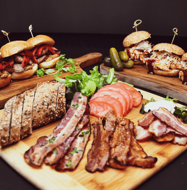 Assorted meats and sandwiches catered on wooden boards, featuring slices of steak, bacon, and garnished with salad and pickles.