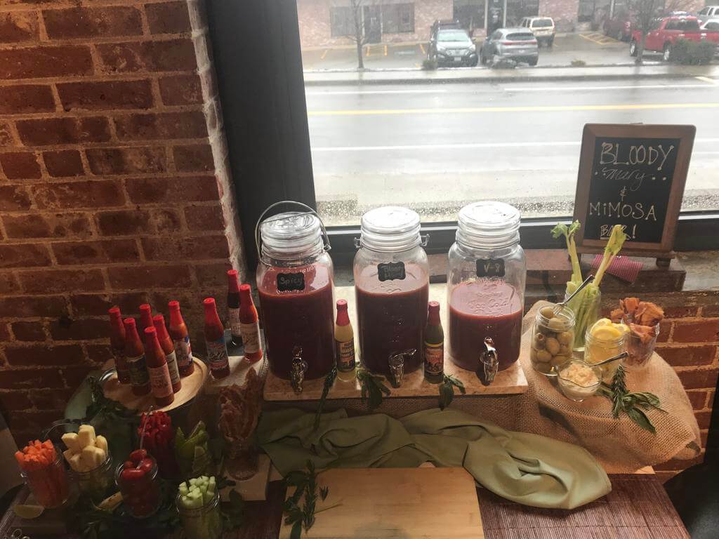 A bloody mary and mimosa bar with various condiments and garnishes on a wooden window ledge.