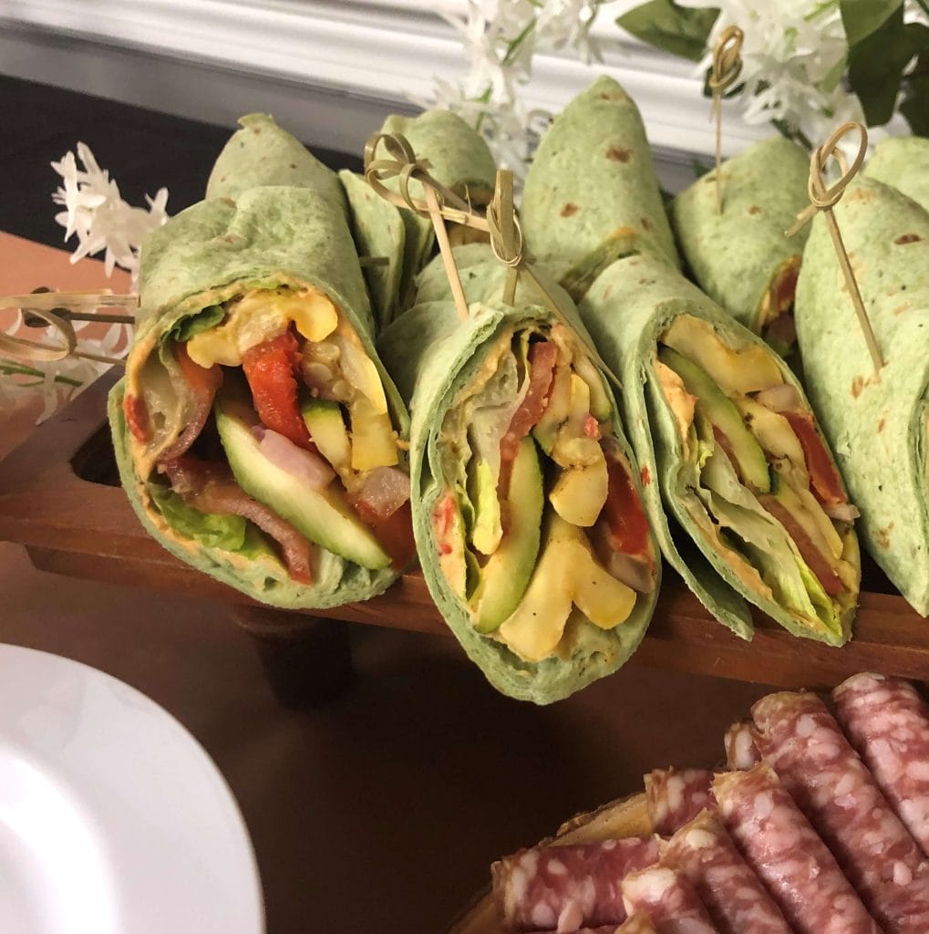 A wooden platter of rolled green tortilla wraps filled with vegetables and chicken, secured with toothpicks, on a wedding buffet table.