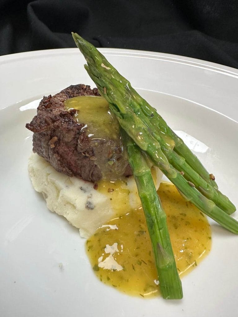 Grilled steak on mashed potatoes with asparagus and sauce, served on a white plate at a wedding bar.