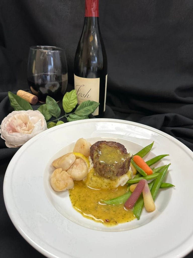 Gourmet meal catered for a wedding with steak and scallops on a white plate, vegetables on the side, accompanied by a glass and bottle of red wine, set against a black backdrop.