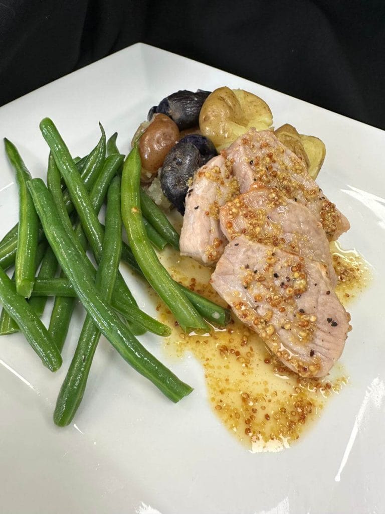 Sliced pork tenderloin with mustard sauce, catered with green beans and roasted potatoes on a white plate.