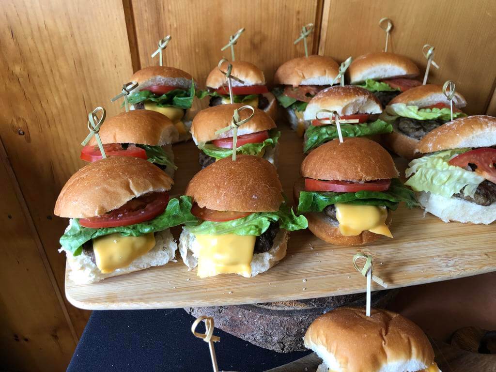 A wooden tray of assorted mini burgers with cheese, lettuce, and tomato.