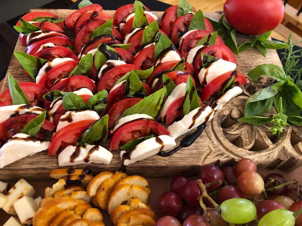 Wooden platter with caprese salad, fresh basil, sliced cheese, crackers, and grapes.