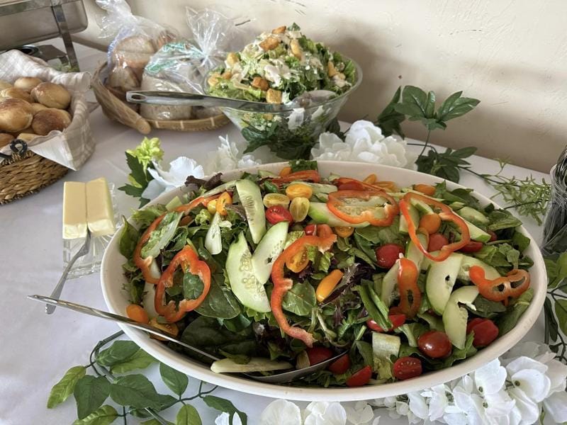 A fresh garden salad served in a large bowl, accompanied by a Caesar salad and bread rolls, on a buffet table adorned with white flowers.