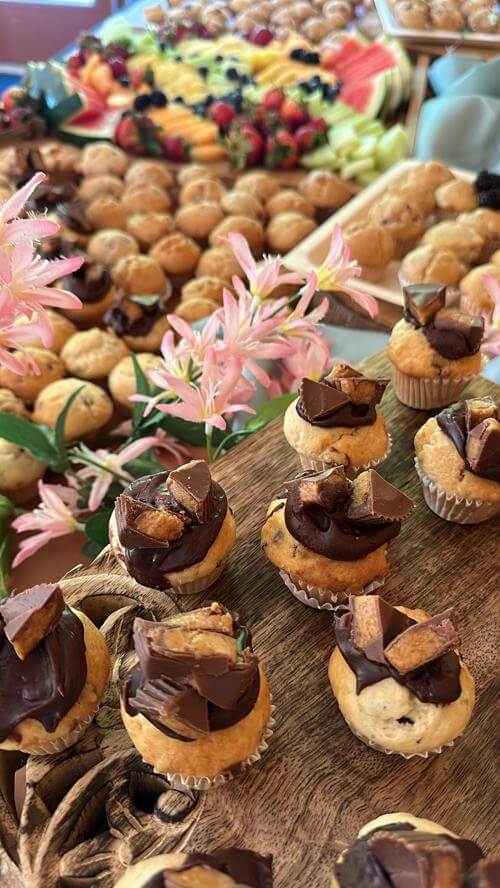 A buffet table filled with assorted pastries and a fruit platter, highlighted by chocolate-topped cupcakes in the foreground and pink flowers for decoration.
