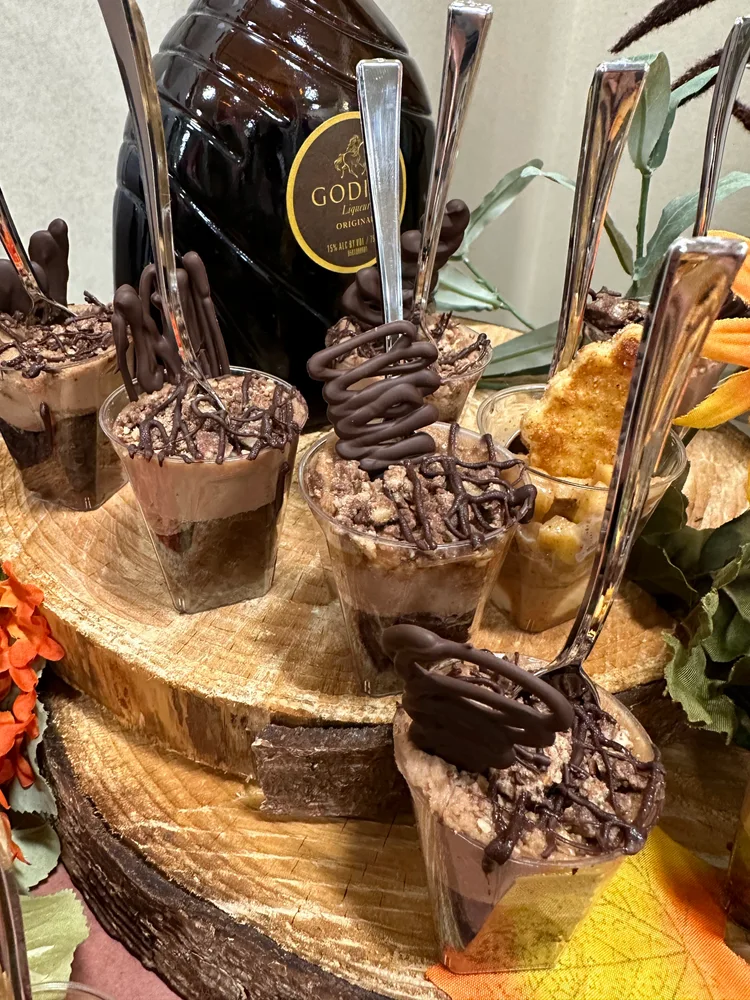 Dessert cups with chocolate and crisped rice toppings, displayed on rustic wooden slices, with a decorative bottle and autumn leaves in the background.