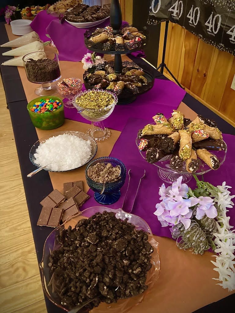 A buffet table with various toppings and chocolate-covered confections, decorated with purple flowers.