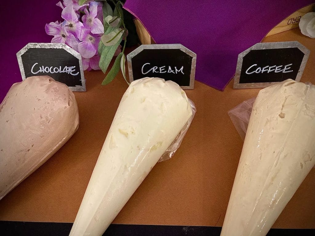 Three cones filled with chocolate, cream, and coffee flavored ice cream, each labeled with small chalkboard signs, displayed on a brown surface with purple flowers in the background.