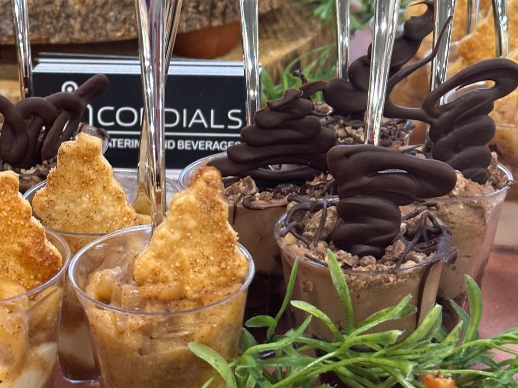 Assorted gourmet desserts in small cups, featuring chocolate-covered pretzels and crispy treats, displayed at a catering event.