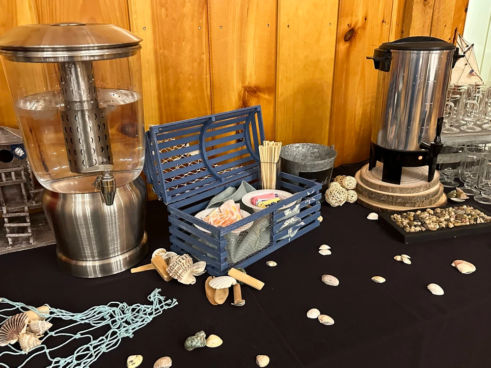 Beverage dispensers, one with water and one with coffee, beside a decorative blue crate with napkins and stirrers, set against a rustic wood backdrop adorned with sea shells and a net.