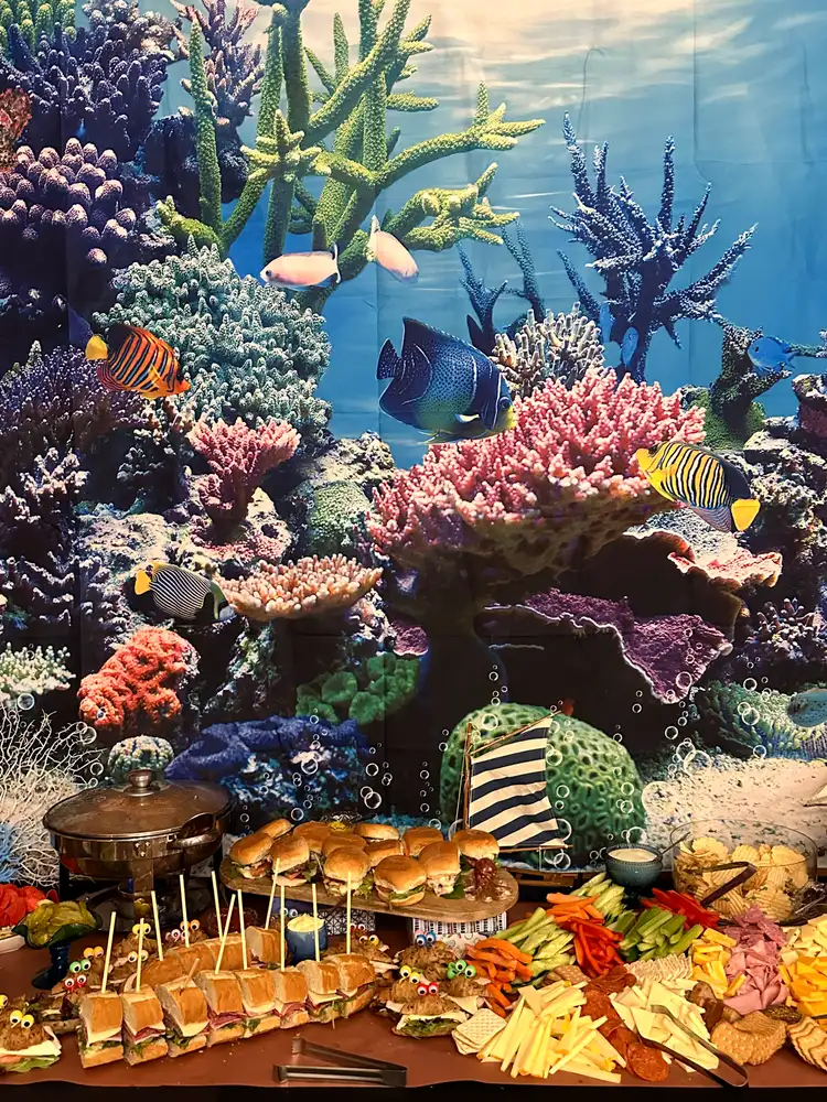Colorful underwater-themed backdrop with a variety of fish and coral, in front of which is a buffet table with sandwiches, cheeses, and dips.