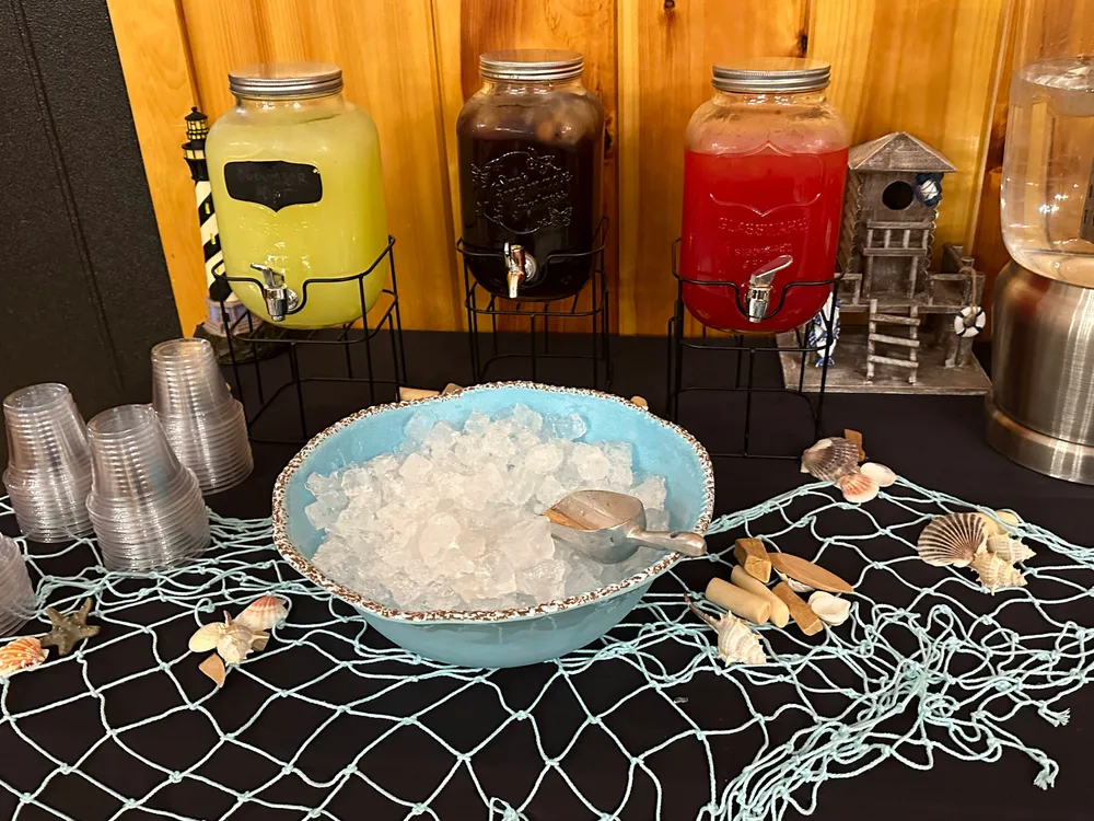 Three large beverage dispensers with yellow, black, and red drinks on a table decorated with shells, a net, and a small lighthouse model, surrounded by ice and cups.