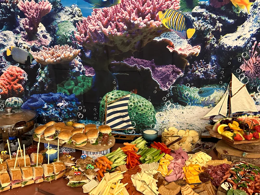 A vibrant buffet with an underwater coral reef backdrop, featuring sandwiches, fruit platters, and assorted appetizers.