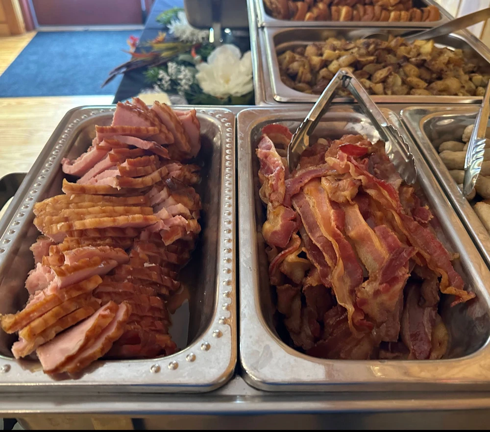 Metal trays containing slices of cooked bacon and ham on a buffet table.
