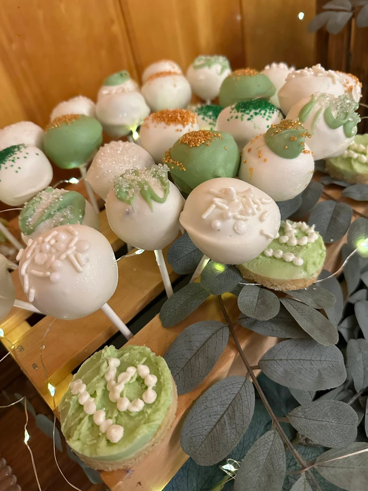 An assortment of green and white cake pops decorated with sprinkles and icing, displayed among eucalyptus leaves on a wooden surface.