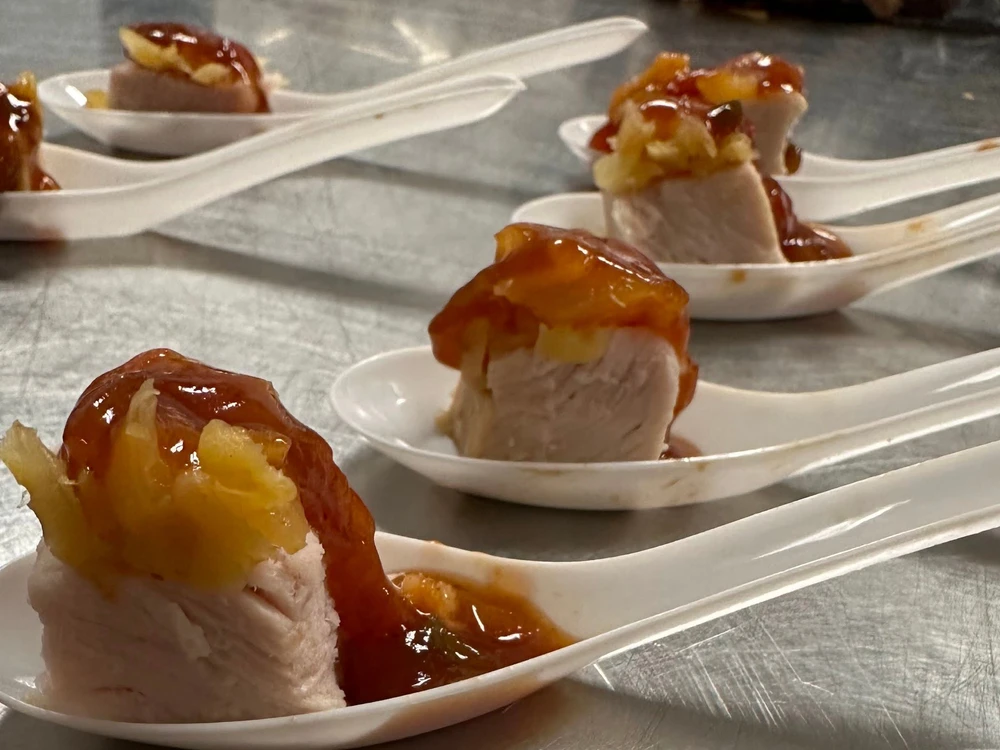 Four ceramic spoons on a metal surface, each holding a piece of pork topped with a golden-brown glaze, likely caramelized sauce.