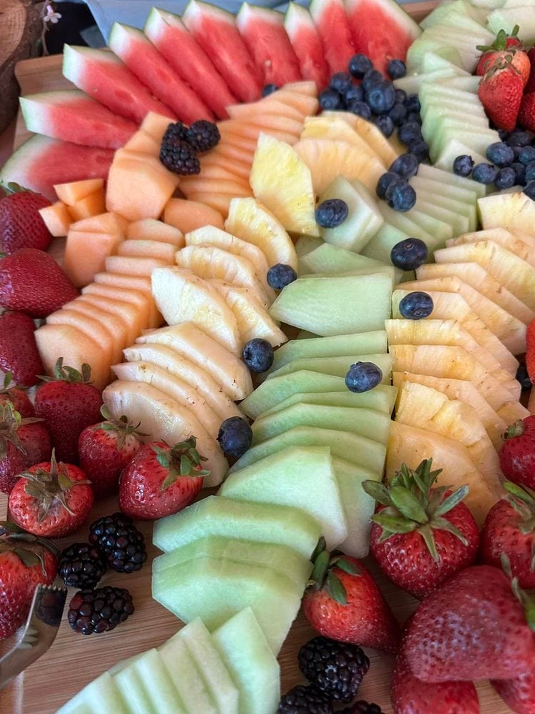 Assorted fresh fruits including strawberries, blueberries, blackberries, watermelon, cantaloupe, pineapple, and honeydew melon artfully arranged on a wooden board.