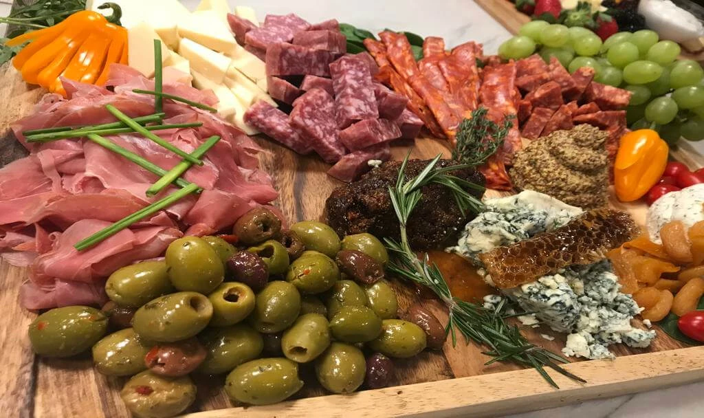 A charcuterie board with assorted meats, cheeses, green and black olives, honeycomb, nuts, cherry tomatoes, grapes, figs, and herbs.