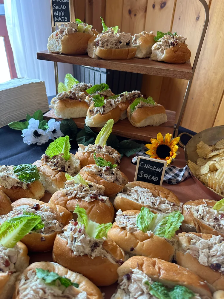Two wooden shelves display chicken salad sandwiches with lettuce. A small chalkboard sign reads "Chicken Salad" and another mentions “Spinach Salad with Walnuts.” Sunflower decorations and potato chips are visible.