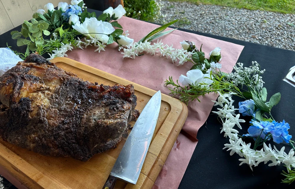 A cooked piece of meat on a wooden cutting board with a knife, surrounded by a floral arrangement of white and blue flowers on a table covered with brown paper.