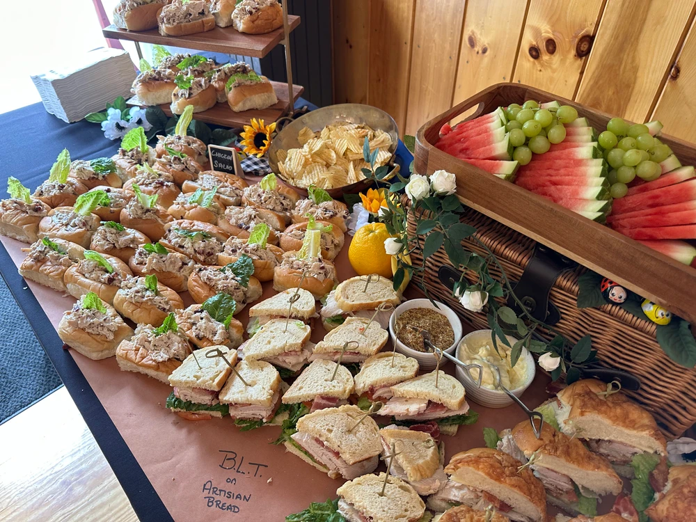 A table is set with a variety of sandwiches, a basket of watermelon and grapes, chips, and small bowls of condiments. A small lemon and floral decoration are also present.
