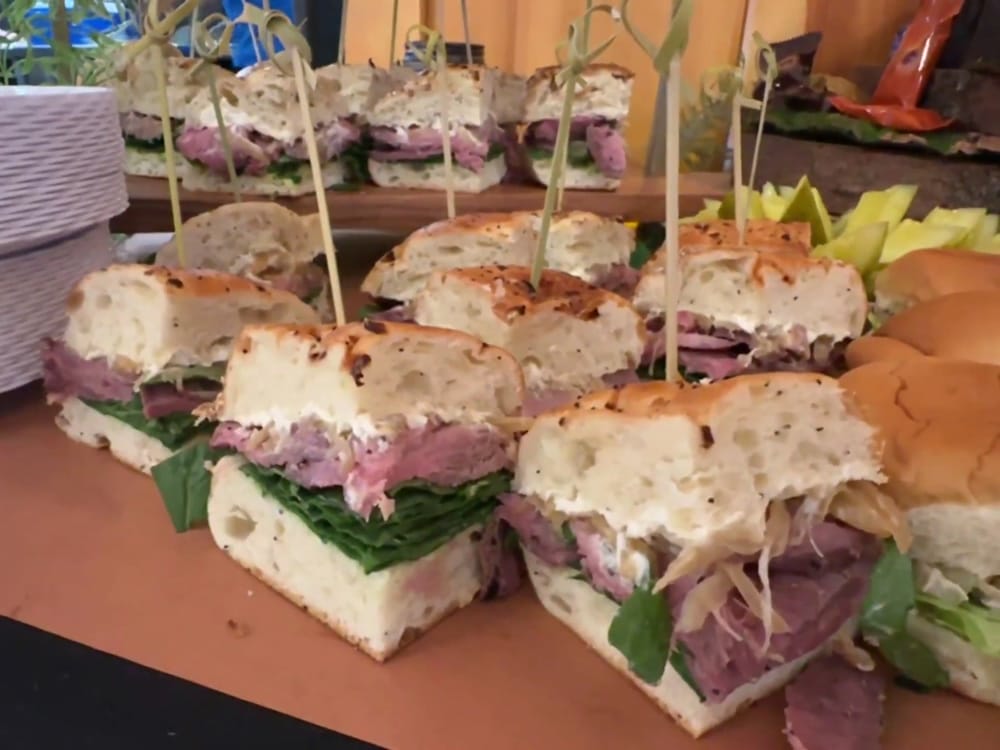 A platter of sandwiches with thick slices of meat, lettuce, and cheese on various types of bread, each held together by decorative skewers.