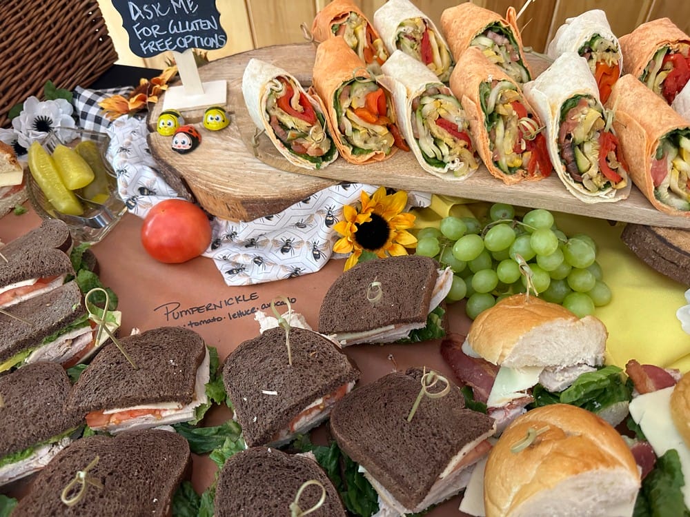 A variety of sandwiches and wraps arranged on a table, including pumpernickel, white bread, and tortilla wraps filled with vegetables. A small sign indicates gluten-free options are available.