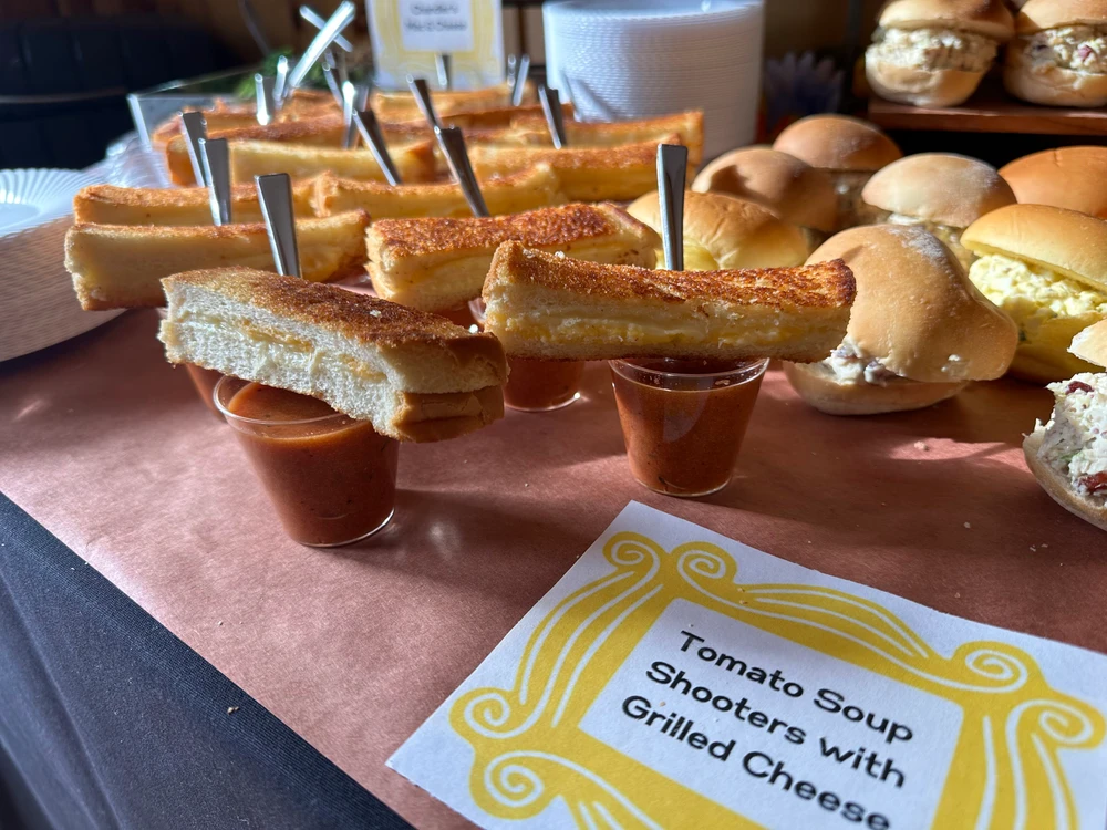 Small glasses of tomato soup topped with grilled cheese sticks, labeled "Tomato Soup Shooters with Grilled Cheese" on a wooden countertop next to an assortment of sliders.