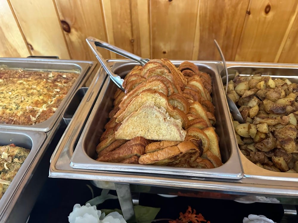 A buffet tray filled with toasted bread slices, flanked by a tray of seasoned potatoes on one side and a tray with a baked egg dish on the other.