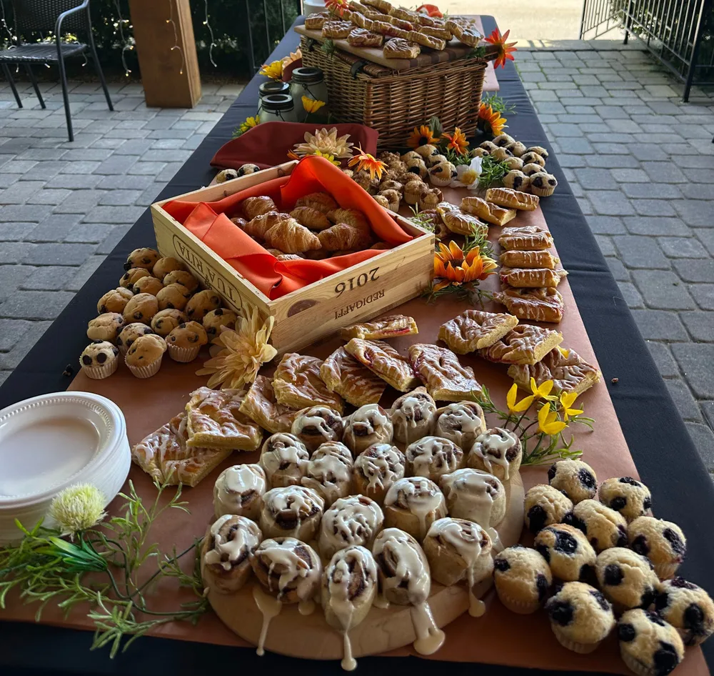 A table is filled with an assortment of pastries, including croissants, muffins, danishes, and glazed buns, all arranged in a decorative manner with flowers and greenery.