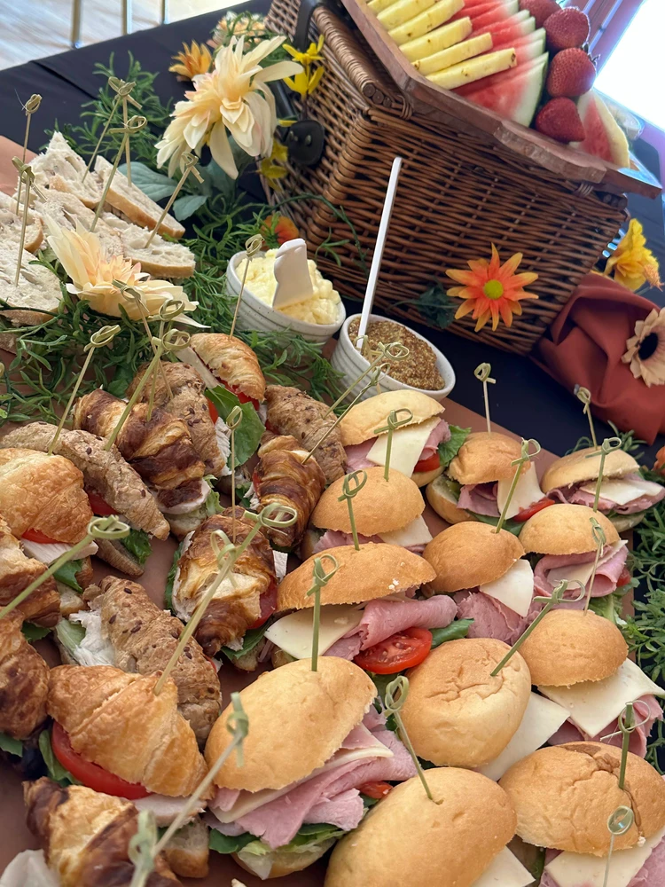 A variety of sandwiches and croissants with various fillings are arranged on a table with decorative flowers. A picnic basket filled with sliced watermelon and other fruits is in the background.