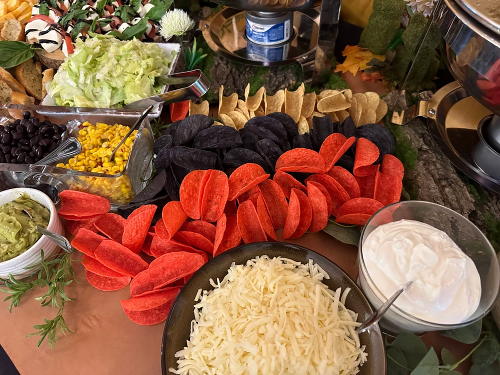 A buffet table with red and blue tortilla chips, grated cheese, sour cream, guacamole, corn, black beans, and chopped lettuce. Various dipping containers and decorations surround the items.