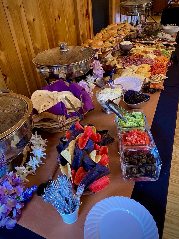 A buffet table with various foods including sandwiches, tortillas, colorful nachos, guacamole, black olives, diced tomatoes, and other dishes set against a wooden wall background. Plates and cutlery are placed nearby.
