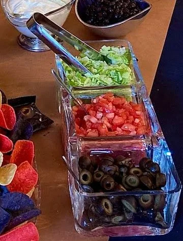 Three clear containers filled with chopped lettuce, diced tomatoes, and sliced black olives on a table with a serving utensil. Nearby, a bowl holds black beans and tortilla chips are visible.
