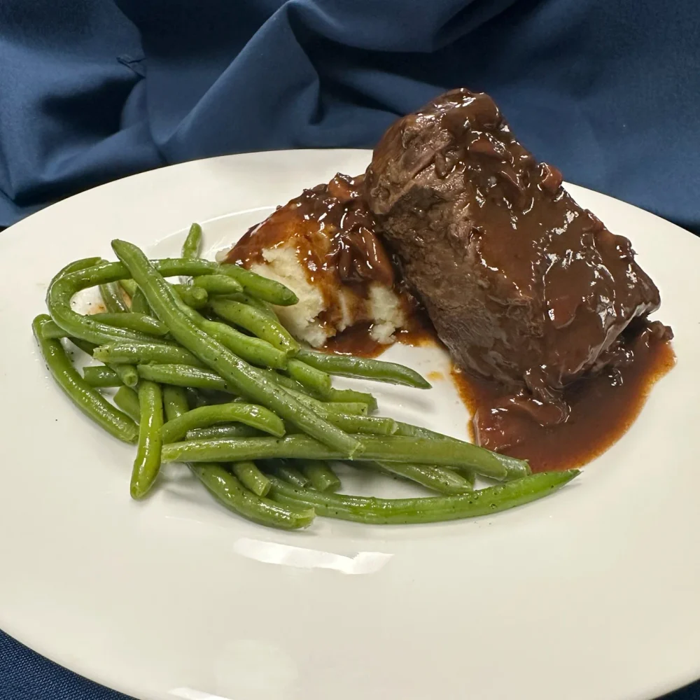 A plate with green beans, mashed potatoes, and a piece of beef covered in brown gravy, set against a blue fabric background.