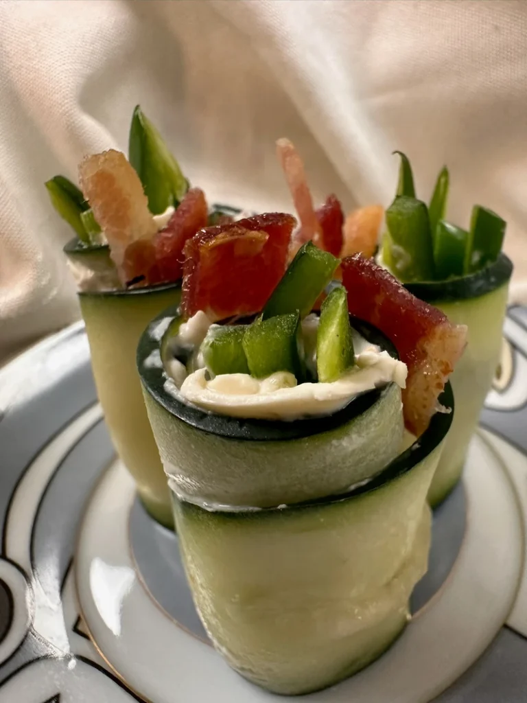 Three cucumber rolls filled with cream cheese, green pepper strips, and pieces of bacon are arranged on a decorative plate.