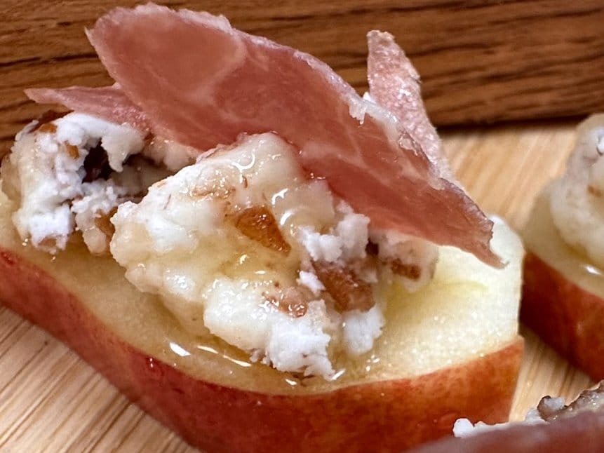 A slice of red pear topped with crumbled blue cheese, a drizzle of honey, and a piece of cured meat on a wooden surface.