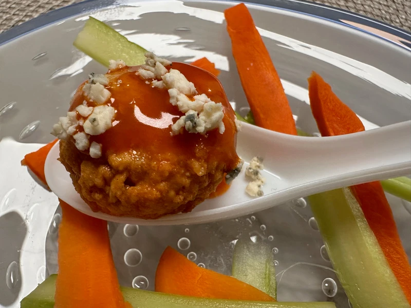 A buffalo chicken meatball with blue cheese crumbles sits on a spoon, surrounded by sliced celery and carrot sticks on a clear plate.