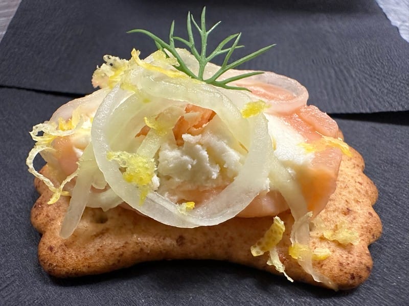 A cracker topped with smoked salmon, cream cheese, sliced onion, lemon zest, and a small sprig of dill, served on a dark napkin.