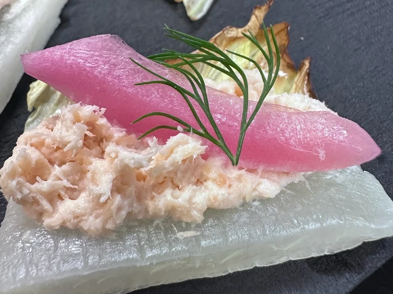 A piece of white vegetable topped with a creamy shredded mixture, a slice of pickled pink radish, and a sprig of fresh dill on a dark surface.