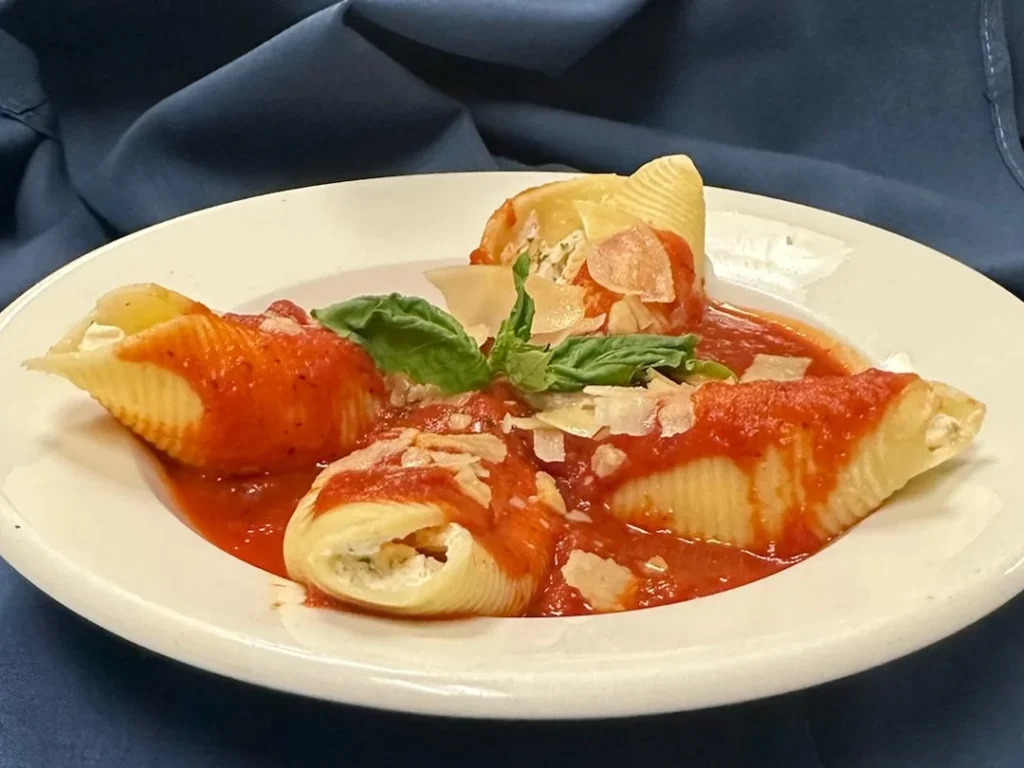 Four stuffed pasta shells with ricotta cheese are topped with tomato sauce, shaved parmesan, and a basil garnish, served on a white plate.