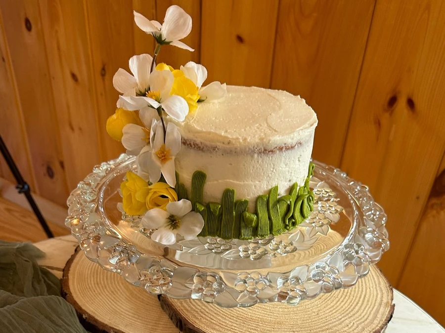 A small round cake with white frosting, green icing decoration at the base, and white and yellow flowers on top, displayed on a glass cake stand.