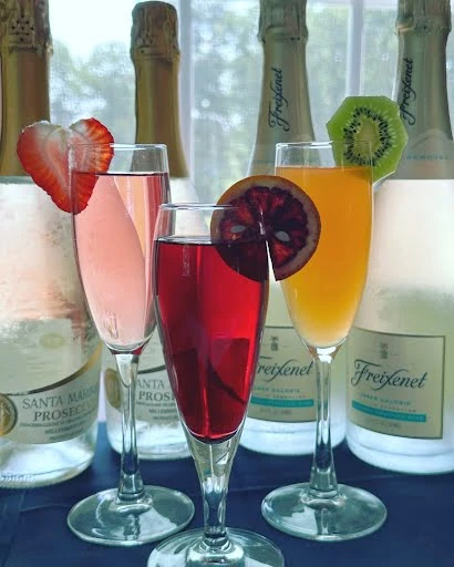 Three glasses of colorful cocktails garnished with fruit slices are in front of bottles of Freixenet and Santa Margherita Prosecco.