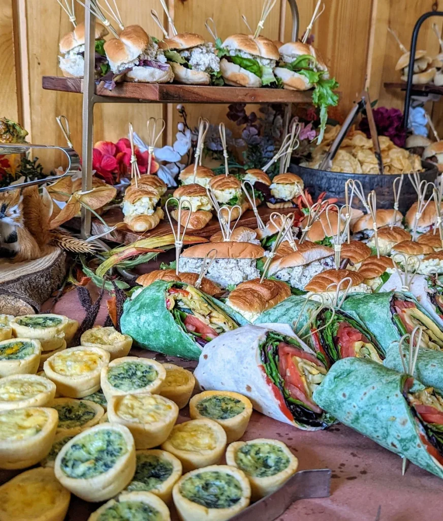 A table displays assorted finger foods including mini sandwiches, wraps, and spinach quiches, arranged on trays and stands with decorative flowers in the background.