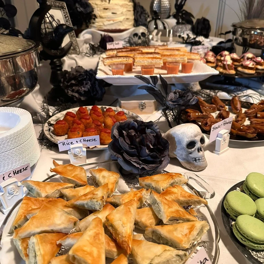 A table set with various foods, pastries, and desserts, decorated with fake skulls, black flowers, and Halloween-themed props.