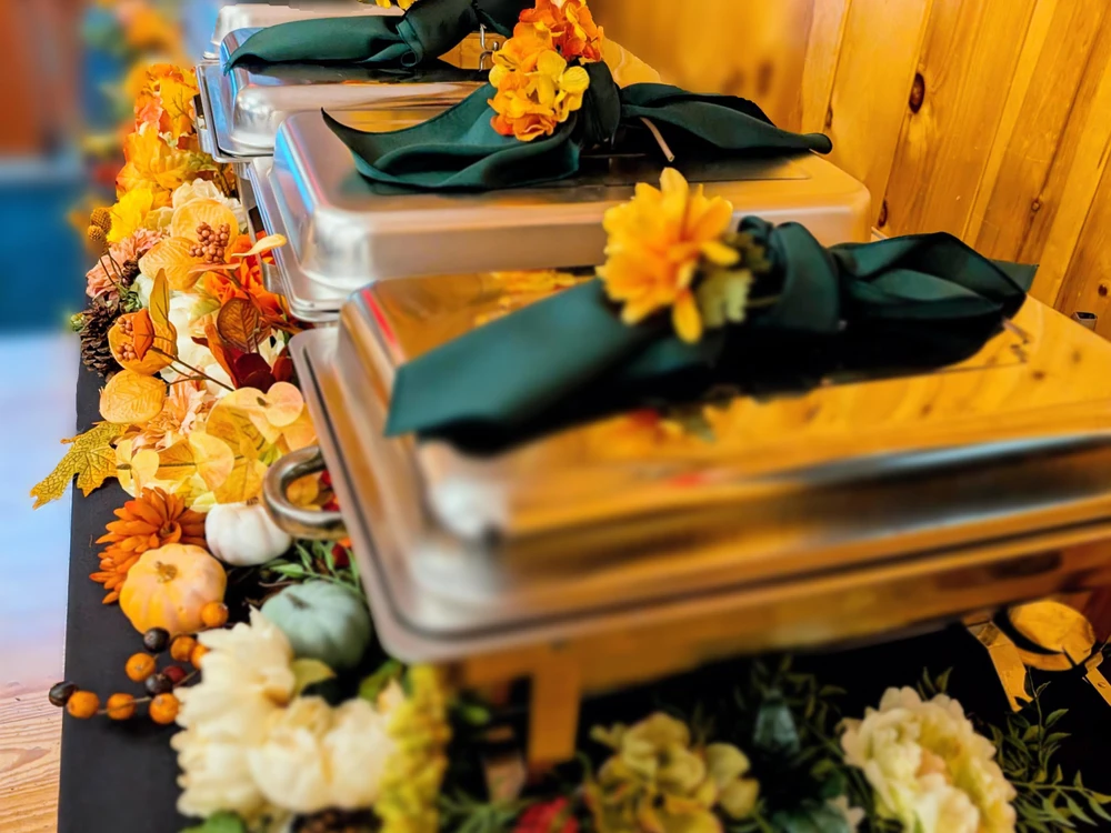Three closed chafing dishes with green napkins and orange flowers on top, set on a table decorated with assorted flowers and autumn-themed decor.