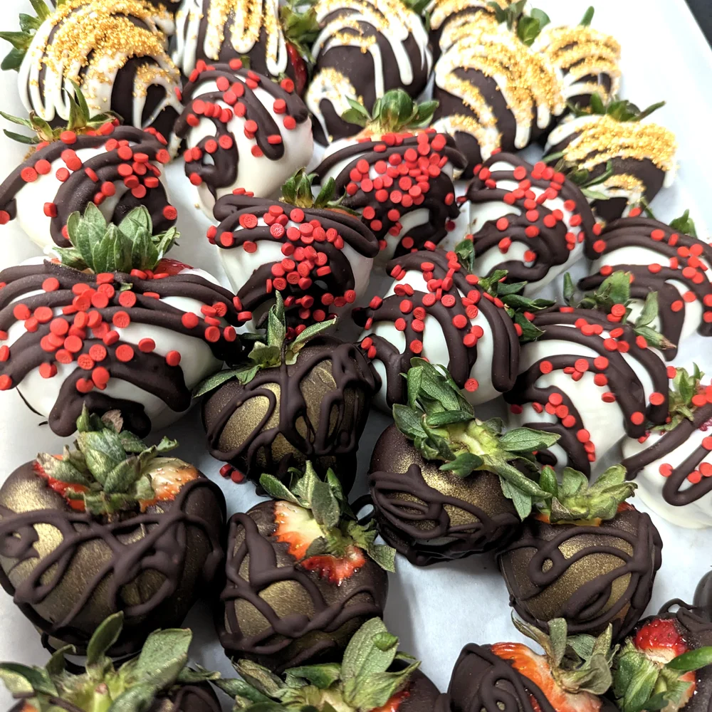 Chocolate-covered strawberries decorated with dark and white chocolate drizzle, red sprinkles, and gold dust, arranged on a white tray.