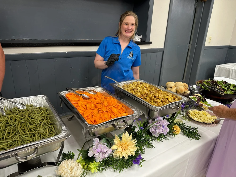 A woman in a blue shirt stands behind a buffet table with green beans, carrots, roasted potatoes, rolls, salad, and garnished flowers.