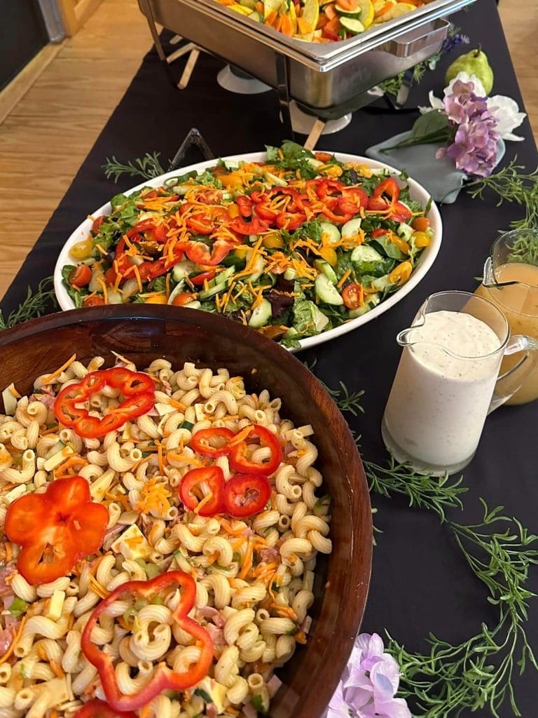 A buffet table with a bowl of pasta salad topped with sliced bell peppers, a mixed green salad, salad dressings, and decorative flowers.
